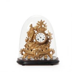 Kaminur af forgyldt metal med skive af hvid emalje, medfølgende glasklokke med base af sortmalet træ. 19. årh.s slutning. H. 30. B. 30. D. 12.