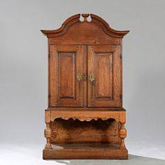 Barok skab af egetræ, svungen top, front med to døre bag hvilke skuffer og rum på senere underdel. 18. årh. H. 132. B. 81. D. 34.